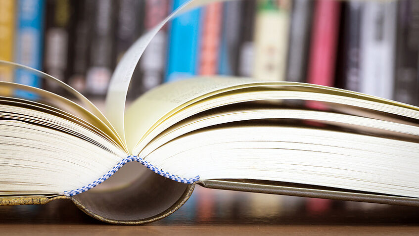 Aufgeschlagenes Buch mit Buchregal im Hintergrund, © iStock.com/pinkomelet