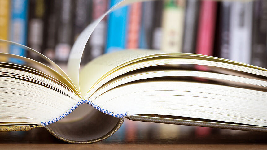 Aufgeschlagenes Buch mit Buchregal im Hintergrund, © iStock.com/pinkomelet